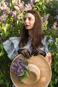 Dreamy young woman looking up, in retro clothes with boatman's straw hat in a blooming lilac garden.