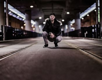 Full length portrait of young man crouching on road below bridge at night