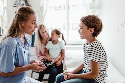 Smiling female pediatrician with otoscope talking to boy while family sitting in background at medical clinic