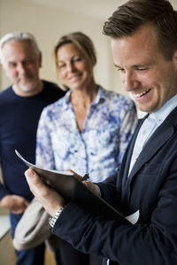 Smiling male real estate agent writing on document with couple standing in background
