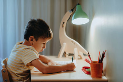 Boy doing his homework while sitting by desk. home interier.diy dinosaur lamp illuminates desk