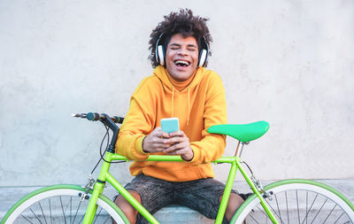 Young man holding smart phone laughing while sitting with bicycle against wall