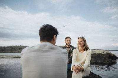 Smiling male and female friends talking to each other while sitting near lake