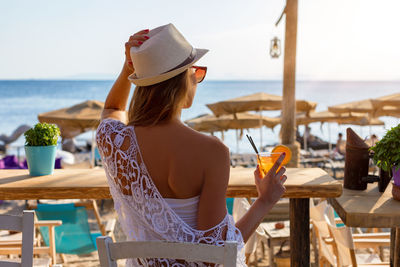 Rear view of woman with drink sitting on chair near beach