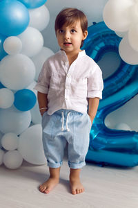 Baby boy in blue pants and shirt standing on the floor in blue and white balloons