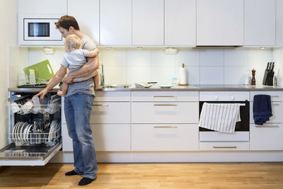 Full length of father working in kitchen while carrying baby at home