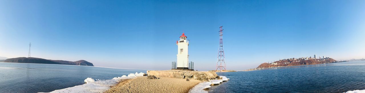 LIGHTHOUSE AMIDST SEA AND BUILDINGS AGAINST CLEAR SKY