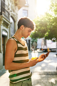 Young male in stylish clothes with takeaway cup of juice browsing cellphone while standing on sunlit city street