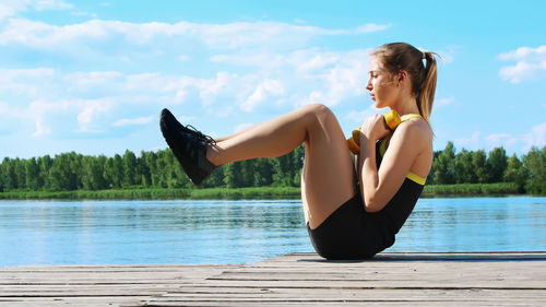 Athletic young blond woman stretching, swings press. river, blue sky and forest in the background
