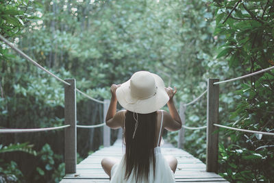 Rear view of woman wearing hat while sitting on bridge in forest