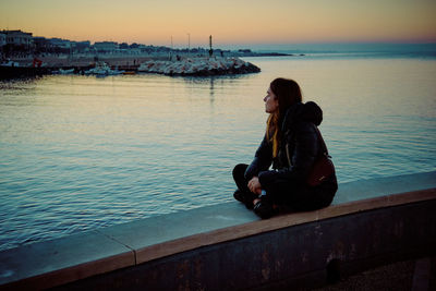 Side view of woman sitting on retaining wall by sea against sky during sunset