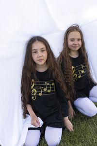 Two little girls, sisters with long hair having fun under sheet on grass in park, making tent