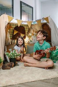 Happy children playing a ukulele and singing on handmade shelter tent in living room at home