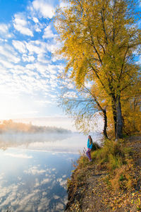 Woman sitting by tree against sky during autumn