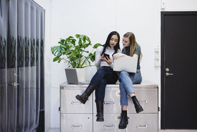 Full length of female bloggers using smart phone while sitting on sideboard in creative office