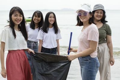 Portrait of smiling friends holding garbage bag while standing at beach against sky