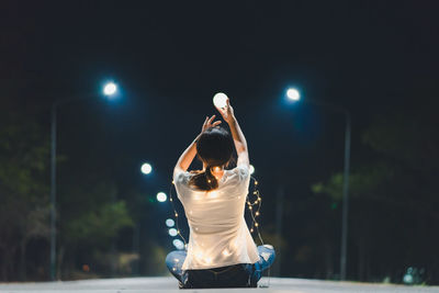 Rear view of woman holding illuminated lighting equipment while sitting on road at night