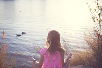 Rear view of girl meditating while sitting by lake