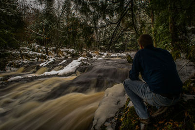 Rear view of man crouching by river in forest during winter