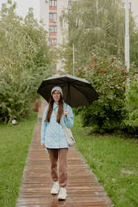 A girl in a blue jacket walks in the park along a wooden path with an umbrella