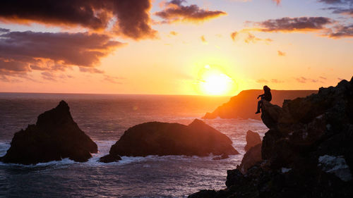 People on rocks by sea against sky during sunset