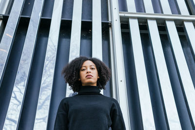 From below serious african american female in black turtleneck looking at camera while standing near metal wall