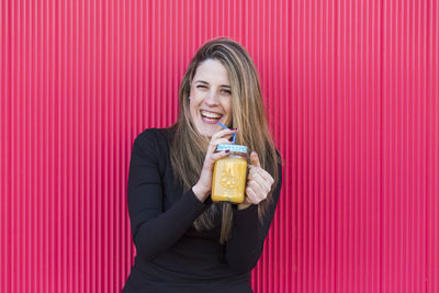 Portrait of young woman drinking juice in glass jar against pink wall