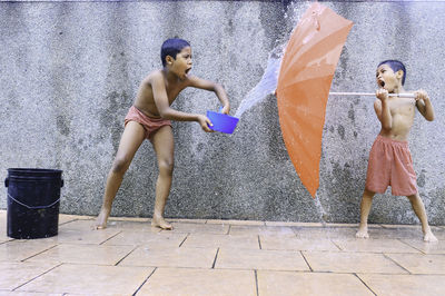 Boy throwing water on brother holding umbrella standing against wall