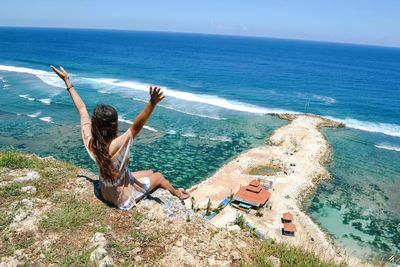 Full length of woman with arms raised sitting on cliff while enjoying sea view
