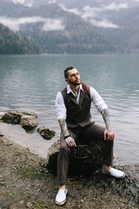 A brutal male hipster groom in a wedding suit in nature by the mountains and lake