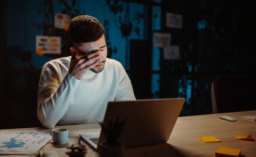 Tired man wearing casual clothes rubbing eyes while working laptop and trying hurry up to deadline