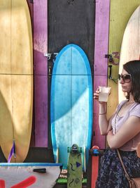Side view of woman having drink while standing by surfboards hanging on wall