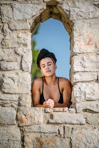 Young woman standing at window amidst stone wall