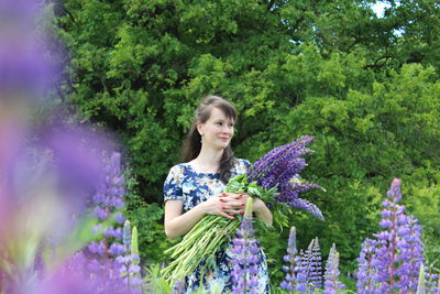 Woman with lavenders looking away while standing on field against trees