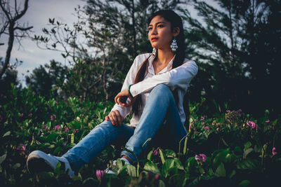 Thoughtful young woman looking away while sitting on field against trees