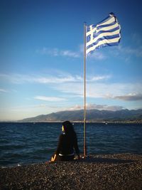 Rear view of woman sitting by greek flag on promenade against sky
