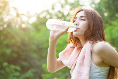 Woman drinking water from bottle against trees