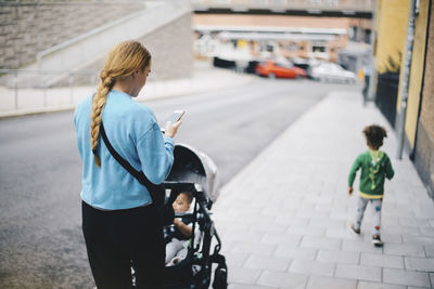 Rear view of mother using mobile phone and pushing baby stroller while walking with son on sidewalk in city