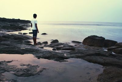 Man standing on rock at sea shore against clear sky