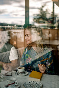 Smiling senior woman sitting with book by male friend in cafe seen through glass