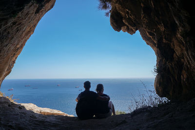 Rear view of couple looking at sea while sitting with arm around against sky