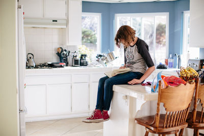 Teen girl sits on her tiled kitchen counter reading a book
