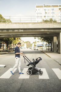 Full length side view of man using smart phone while pushing baby carriage on street in city