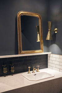 Mirror with golden frame hanging on black wall near lamp over marble counter with sink in elegant bathroom