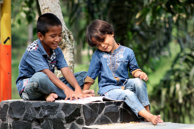 Boys reading book while sitting on seat against tree