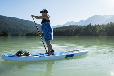 Full body pregnant woman in dress paddleboarding on calm water of lake against mountain ridge and green forest on summer day in british columbia, canada