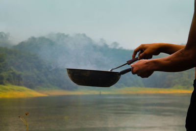 Midsection of man preparing food in kitchen utensil standing by lake