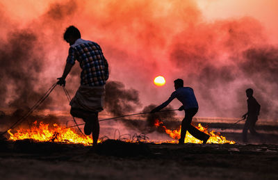 Silhouette people with fire against sky at sunset