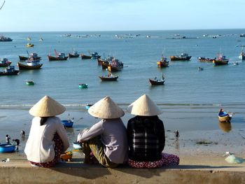 Rear view of women sitting on retaining wall while looking at fishing boats in sea