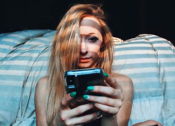 Portrait of young woman with blond hair using mobile phone while sitting by bed at home